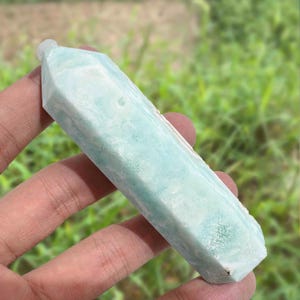 May include: A light blue crystal wand with a faceted, hexagonal shape. The crystal has a slightly rough texture and is held in a hand against a blurred green background.