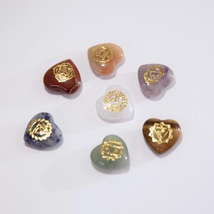May include: Seven heart-shaped stones in various colours, including red, orange, purple, white, brown, blue, and green. Each stone features a gold-coloured symbol. The stones are arranged in a circular pattern on a white surface.