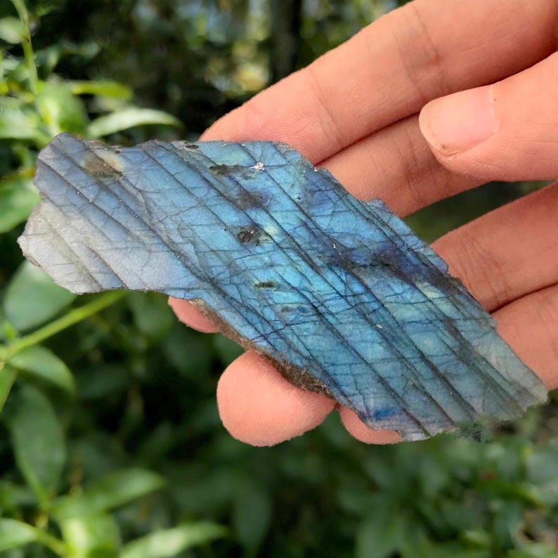 May include: A piece of labradorite stone with a blue and gray iridescent surface. The stone has a rough, natural texture and displays a play of color. The stone is held in a hand against a blurred green background.