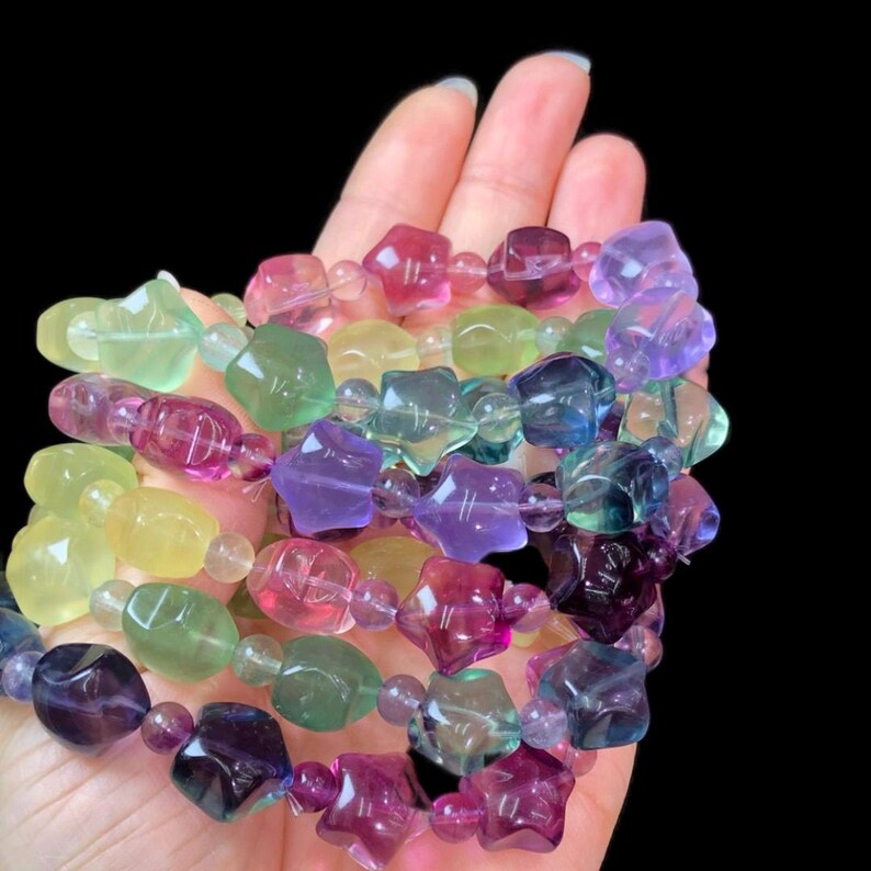 May include: A collection of colorful gemstone bracelets. The beads are various shapes and sizes, including stars and rounded shapes. The colors include shades of purple, pink, green, and yellow. The bracelets are held in a hand against a black background.