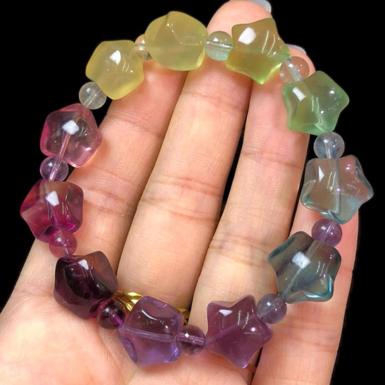 May include: A colorful bracelet featuring star-shaped beads in shades of purple, pink, green, and yellow. The bracelet is strung with smaller, round beads and has a gold-colored clasp. The gemstones are faceted and appear to be polished.