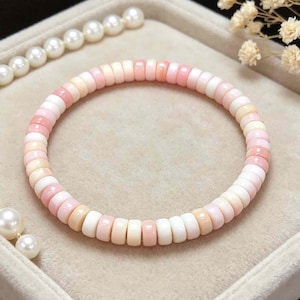Könnte beinhalten: Ein rundes Armband aus kleinen, gleichmäßig großen Perlen in Rosa-, Weiß- und Cremetönen. Das Armband wird auf einer neutralen Oberfläche präsentiert, mit einer Perlenkette und kleinen weißen Blumen im Hintergrund. Schmuck.