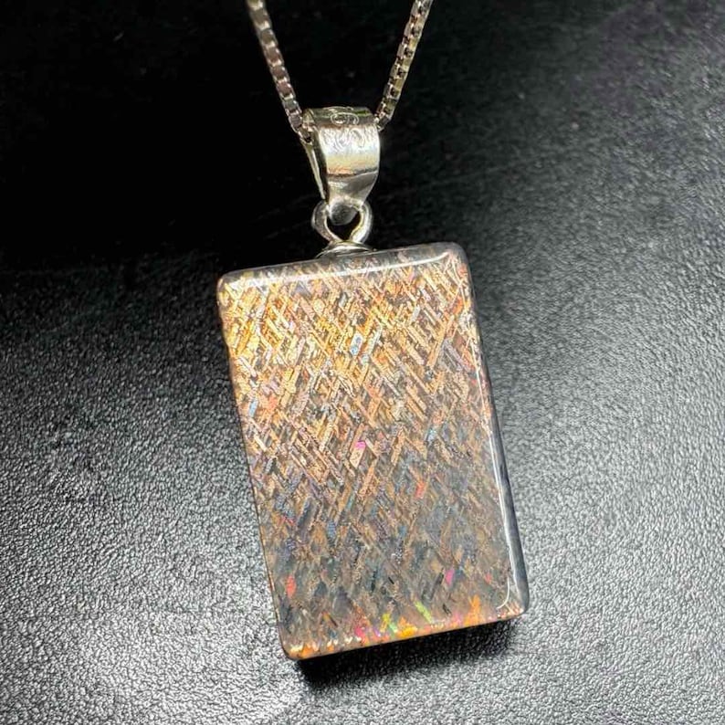 May include: A rectangular pendant with a silver chain. The pendant has a shimmering, multi-colored surface with gold, brown, and iridescent hues. The pendant is approximately 1 inch long.