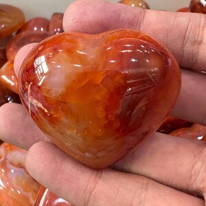 May include: A polished, heart-shaped carnelian stone. The stone displays a mix of orange and reddish-brown hues with a glossy finish. The heart is held in a person's hand, showcasing its smooth, rounded form. This gemstone is a decorative item.