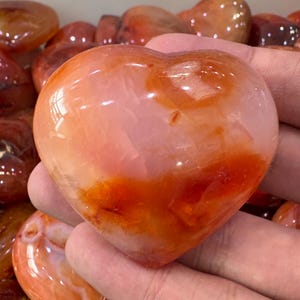 May include: A heart-shaped carnelian stone with a polished, glossy finish. The stone features a blend of translucent pink, orange, and red hues. The heart is held in a hand, with other similar stones visible in the background.