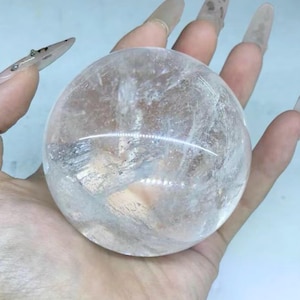 Puede incluir: Una esfera de cristal de cuarzo transparente, sostenida en una mano. La esfera es transparente con inclusiones internas y una superficie lisa y pulida. El fondo es azul claro.