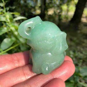 Peut inclure: Petite figurine d'éléphant en aventurine verte. L'éléphant est sculpté avec des détails, notamment une trompe, des oreilles et une expression souriante. La figurine est tenue dans une main, avec un arrière-plan flou de feuillage vert.