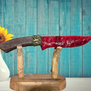 Puede incluir: Un cuchillo decorativo con un mango marrón oscuro y una hoja roja vibrante. El mango está adornado con detalles dorados. El cuchillo descansa sobre un soporte de madera con acabado natural, sobre un fondo azul.