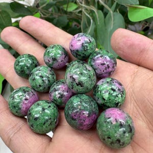 May include: A selection of polished gemstone spheres. The spheres are a mix of green and pink, with a speckled appearance. The spheres are held in an open hand, with a natural background.