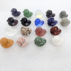 Puede incluir: Una colección de pequeñas figuras de patos talladas en varias piedras preciosas y minerales. Los patos son de varios colores, incluyendo verde, azul y piedra moteada. Los patos están dispuestos sobre una superficie blanca.
