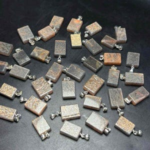 May include: A collection of rectangular pendants with silver-colored bails. The pendants have a variety of textures and colors, including silver, gray, brown, and gold. Some pendants have etched patterns. The pendants are scattered on a dark surface.