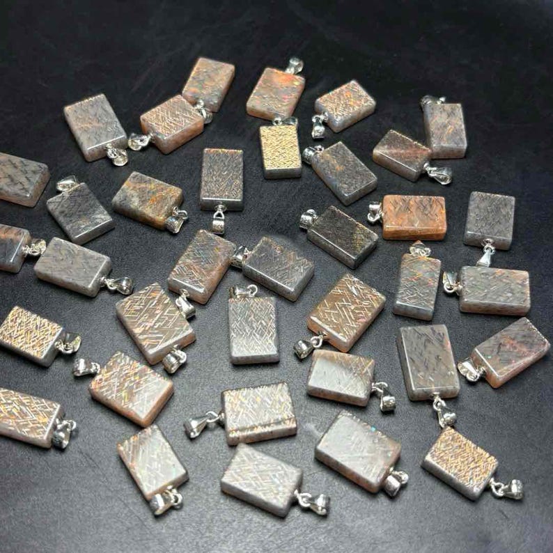 May include: A collection of rectangular pendants with silver-colored bails. The pendants have a variety of textures and colors, including silver, gray, brown, and gold. Some pendants have etched patterns. The pendants are scattered on a dark surface.