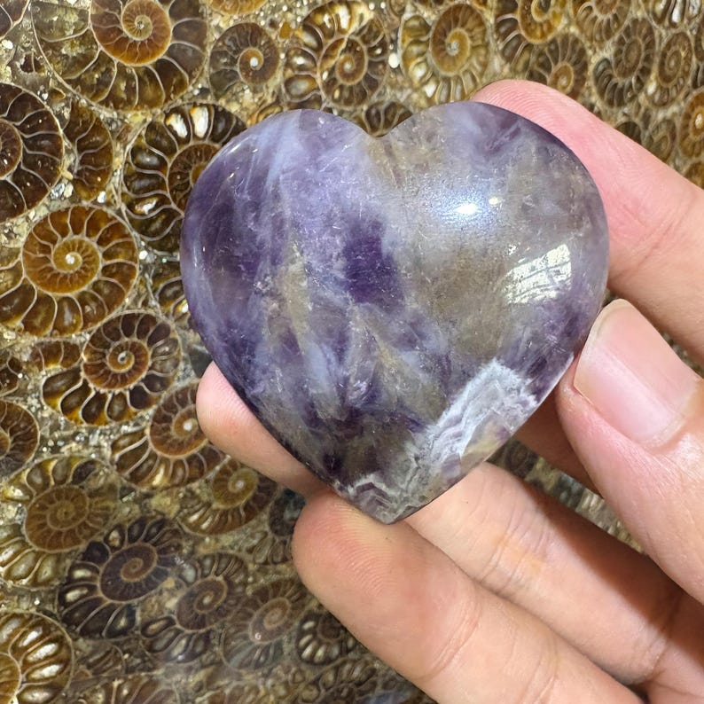 May include: A heart-shaped amethyst crystal with shades of purple, white, and brown. The polished stone has a smooth surface and is held in a hand. The background features a pattern of brown spiral shapes.