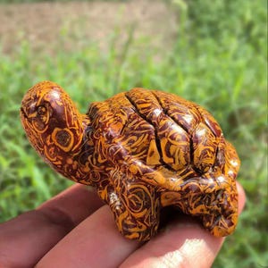 Puede incluir: Figura de tortuga tallada a mano con un diseño de patrón marrón y amarillo. El caparazón y el cuerpo de la tortuga están intrincadamente detallados, mostrando un estilo artístico único. La figura se sostiene en una mano, con un fondo verde borroso.