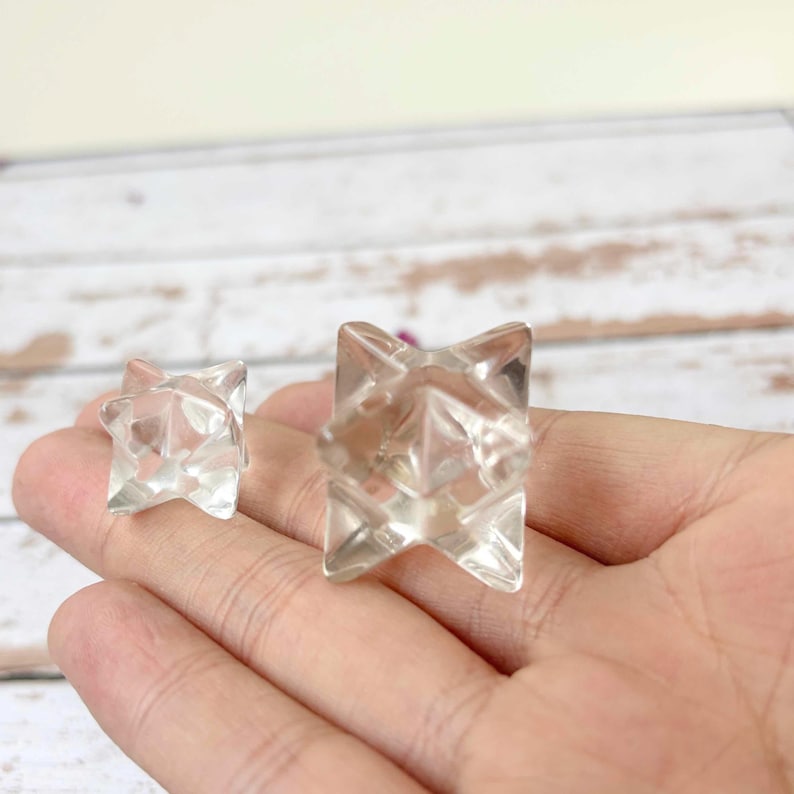 May include: Two clear, faceted crystal star-shaped objects. The objects are transparent and have a geometric design. The larger star is in focus, while the smaller one is slightly blurred. The objects are displayed on a light-colored surface.