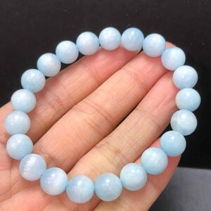 Op de afbeelding: Een lichtblauwe kralenarmband. De armband is gemaakt van ronde, gepolijste kralen die aan elkaar zijn geregen. De kralen hebben een doorschijnende kwaliteit en een zachte pastelkleur. De armband wordt tegen een donkere achtergrond getoond.