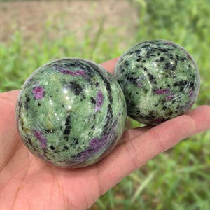 Op de afbeelding: Twee gepolijste edelsteen bollen. De bollen zijn groen met zwarte en paarse insluitsels. De bollen worden in een hand gehouden, met een vage groene achtergrond.