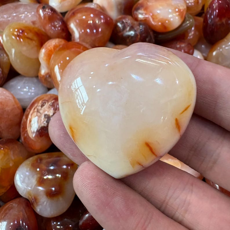 May include: A heart-shaped, polished stone with a creamy white base and orange-red streaks. The stone is held in a hand, with other similar stones in the background. The stones have a smooth, glossy finish.