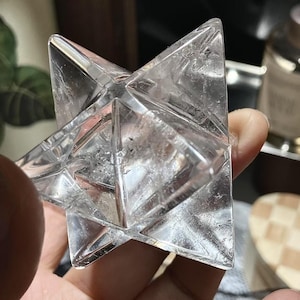 May include: A clear, multi-faceted crystal star, held in a hand. The crystal is transparent with visible internal structures and sharp, geometric edges. The star shape is symmetrical and appears to be made of multiple pyramid shapes joined together.