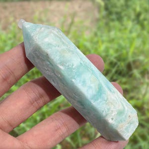 May include: A light blue crystal point with a polished, faceted design. The crystal has a translucent quality with subtle variations in color. The crystal is held in a hand against a blurred green background.