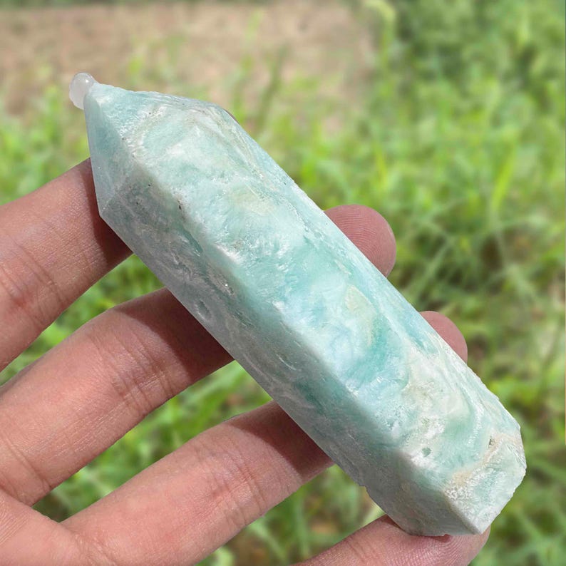 May include: A light blue crystal point with a polished, faceted design. The crystal has a translucent quality with subtle variations in color. The crystal is held in a hand against a blurred green background.