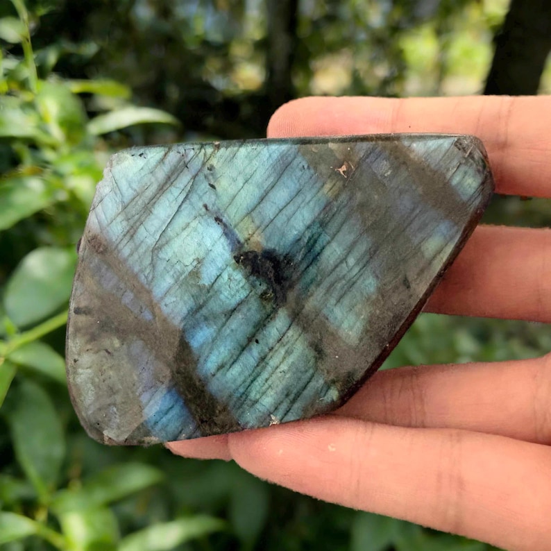 May include: A polished labradorite gemstone with a mix of blue, teal, and gray hues. The stone has a natural, irregular shape and displays iridescent flashes. The surface has visible lines and patterns, adding to its unique appearance.