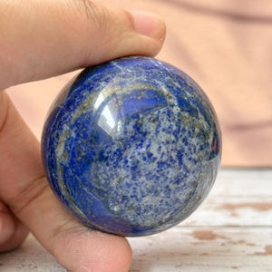 以下が含まれることがあります： 研磨されたラピスラズリの球体。金と灰色のインクルージョンを持つ濃い青色の宝石。球形の石は光を反射し、手で持たれています。装飾品は滑らかで丸みを帯びた球体です。