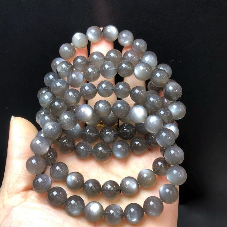May include: Multiple strands of grey moonstone bead bracelets. The beads are round and have a subtle sheen, with variations in colour from light grey to darker shades. The bracelets are displayed against a dark background.
