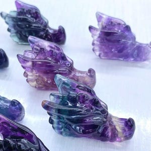 May include: Several carved dragon heads are displayed, each in shades of purple, green, and clear. The dragon heads are made of a translucent material, with detailed features including wings and scales. The heads are arranged on a white surface.