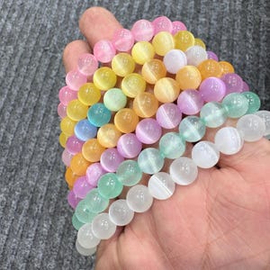 May include: A collection of colorful beaded bracelets in shades of pink, yellow, green, blue, orange, and purple. The bracelets are made of round, polished beads, and are held in a hand. The beads have a cat's eye effect.