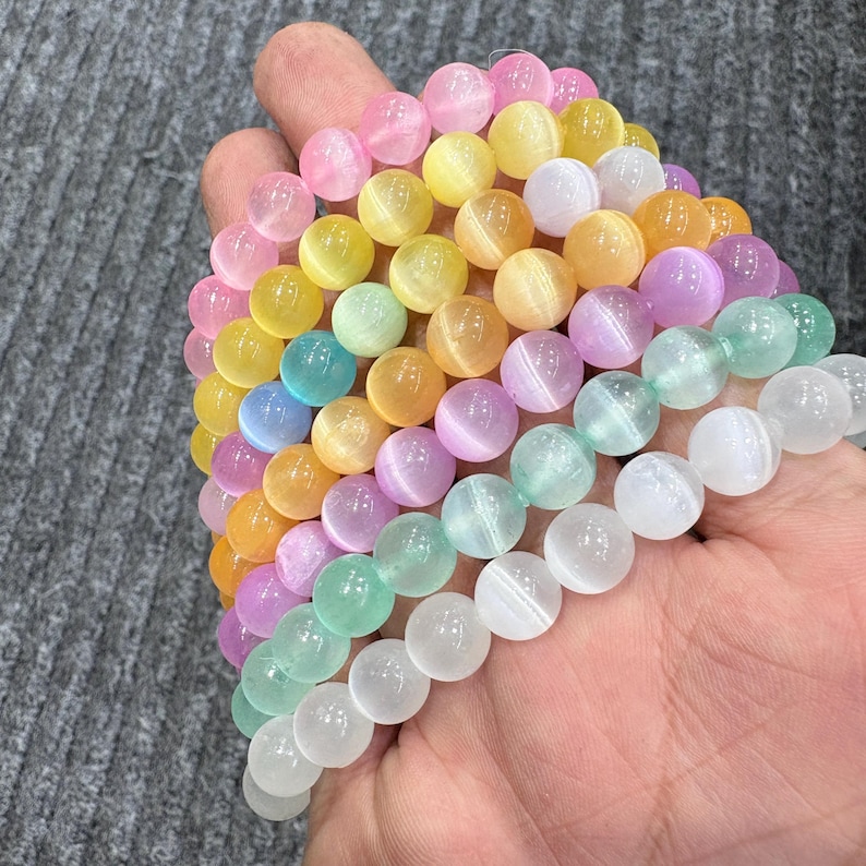 May include: A collection of colorful beaded bracelets in shades of pink, yellow, green, blue, orange, and purple. The bracelets are made of round, polished beads, and are held in a hand. The beads have a cat's eye effect.