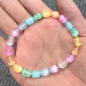 May include: A colorful bracelet featuring a string of translucent, barrel-shaped beads in a rainbow of pastel colors. The beads are light pink, yellow, orange, green, and blue. The bracelet is shown on a hand.