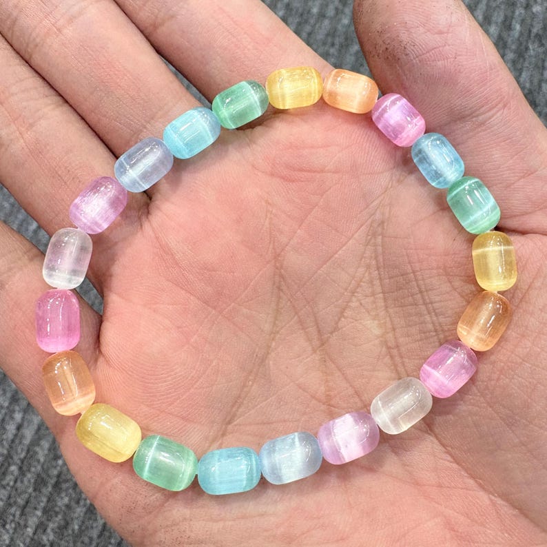 May include: A colorful bracelet featuring a string of translucent, barrel-shaped beads in a rainbow of pastel colors. The beads are light pink, yellow, orange, green, and blue. The bracelet is shown on a hand.