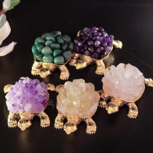 Könnte beinhalten: Fünf dekorative Schildkrötenfiguren. Jede Schildkröte hat eine goldfarbene Metallbasis und Beine. Die Panzer bestehen aus verschiedenen farbigen Edelsteinen, darunter grüne, violette und klare Kristalle. Die Schildkröten sind auf einer dunklen Oberfläche angeordnet.
