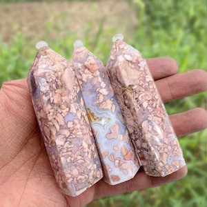 May include: Three polished gemstone obelisks with a mix of peach, cream, and lavender colors. Each obelisk has a clear, rounded top. The stones are held in a hand against a blurred green background.