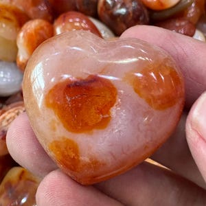 May include: A heart-shaped stone with a polished surface. The stone is primarily a pale pink color with orange and brown spots. The stone is smooth and appears to be a decorative item.