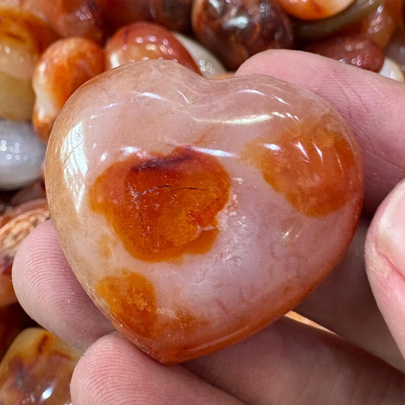 May include: A heart-shaped stone with a polished surface. The stone is primarily a pale pink color with orange and brown spots. The stone is smooth and appears to be a decorative item.