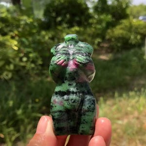 May include: A carved stone torso figurine, displaying green, black, and pink colours. The figure's arms are positioned across the chest. The sculpture is held in a hand, with a blurred green background.