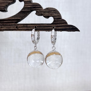 May include: A pair of silver hoop earrings with clear, spherical pendants. The hoops are adorned with small, sparkling stones. Each pendant has a gold-colored band around the middle. The earrings are displayed on a dark wooden jewelry holder.