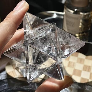 May include: A clear, multi-faceted crystal star held between fingers. The star has a complex geometric design with sharp angles and a slightly cloudy interior. The crystal is transparent, allowing light to pass through, and appears to be a decorative item.