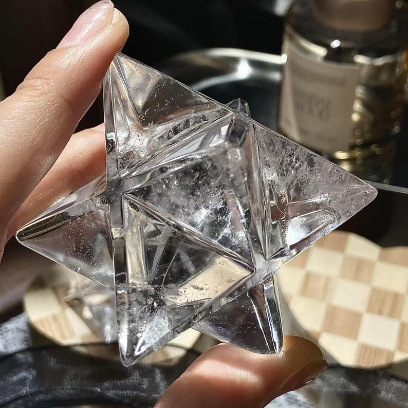 May include: A clear, multi-faceted crystal star held between fingers. The star has a complex geometric design with sharp angles and a slightly cloudy interior. The crystal is transparent, allowing light to pass through, and appears to be a decorative item.