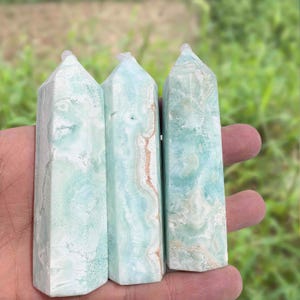 May include: Three light blue crystal obelisks with white and brown veining are held in a hand. The obelisks have a hexagonal shape and a pointed top. The background is blurred green.