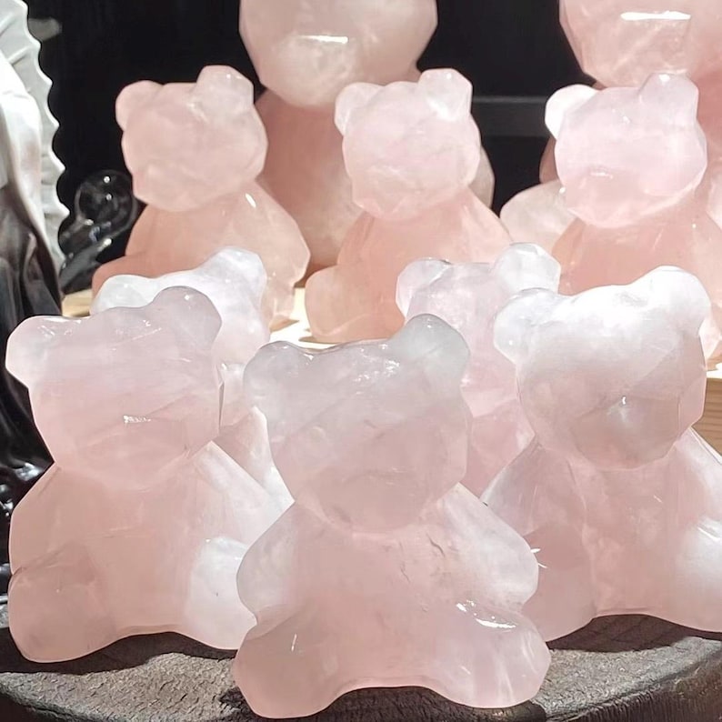 May include: A collection of rose quartz bear figurines in various sizes. The bears are a soft pink color and have a smooth, polished finish. The figurines are arranged together, showcasing their carved details.