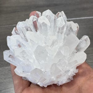 May include: A cluster of clear quartz crystals, showcasing a multitude of pointed formations. The crystals are translucent, reflecting light and creating a sparkling effect. The quartz cluster is held in a hand, highlighting its size and natural beauty.