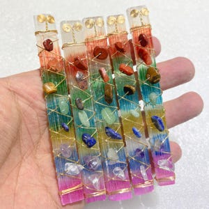 May include: Five rectangular selenite wands featuring a vertical rainbow gradient of red, orange, yellow, green, blue, and purple. Each wand is wrapped in gold-colored wire and adorned with seven small, polished, multi-colored gemstone chips.