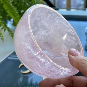 Puede incluir: Un cuenco de cuarzo rosa translúcido, de color rosa pálido, con un borde pulido. El cuenco se sostiene en una mano, mostrando su textura cristalina natural. El interior del cuenco es visible, revelando su color rosa claro y sus patrones únicos.