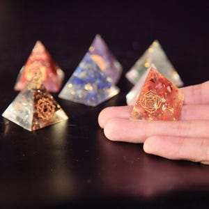 Puede incluir: Una colección de pequeñas esculturas de resina en forma de pirámide, coloridas. Una se sostiene en una mano, mostrando un símbolo de chakra dorado. Las pirámides presentan piedras incrustadas y escamas metálicas en rojo, azul, verde y dorado. El fondo es oscuro.