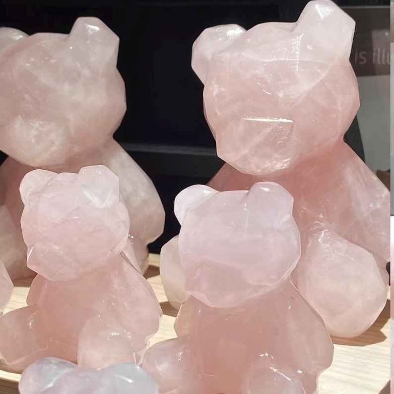 May include: Several pink rose quartz bear figurines of varying sizes are displayed. The bears have a faceted, geometric design, and are a soft, translucent pink color. The background is dark, highlighting the sculptures.
