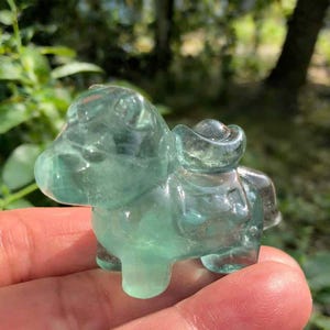 Può includere: Una statuetta di cristallo verde chiaro traslucido di un piccolo animale, forse un cavallo o un asino, con una struttura a forma di sella sulla schiena. La statuetta è tenuta in mano, con uno sfondo verde sfocato.