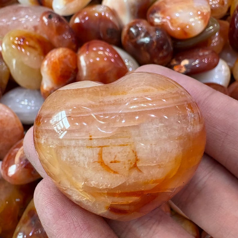 May include: A heart-shaped, polished carnelian stone with bands of orange and white. The stone is held in a hand, with other tumbled stones in the background. The carnelian has a glossy finish.